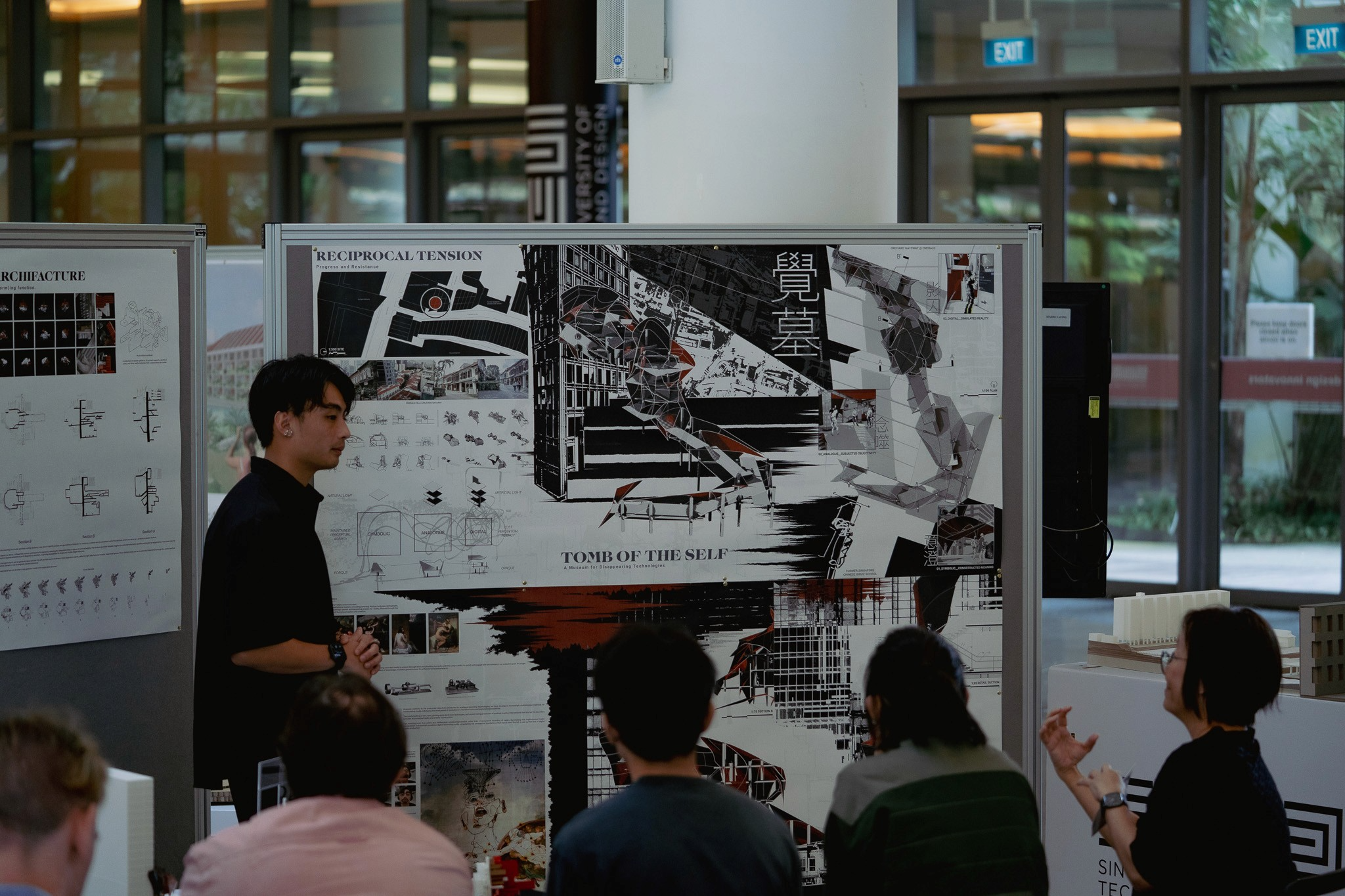 Person presenting an architectural project board titled "Tomb of the Self" to a seated audience in a bright exhibition space.